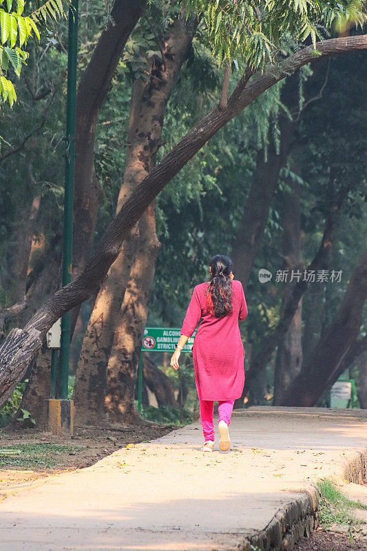 图片无法识别，印度妇女穿着亮粉色，Salwar Kameez传统服装，沿着公共公园的步道走到远处，同时打电话，后视图